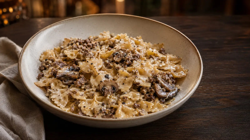 A bowl of creamy pasta with ground meat and mushrooms, served on a dark wooden table with a folded napkin.