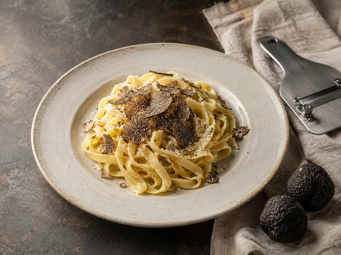 Tagliatelle with black truffle shavings, whole truffles and a truffle slicer on a stone worktop.